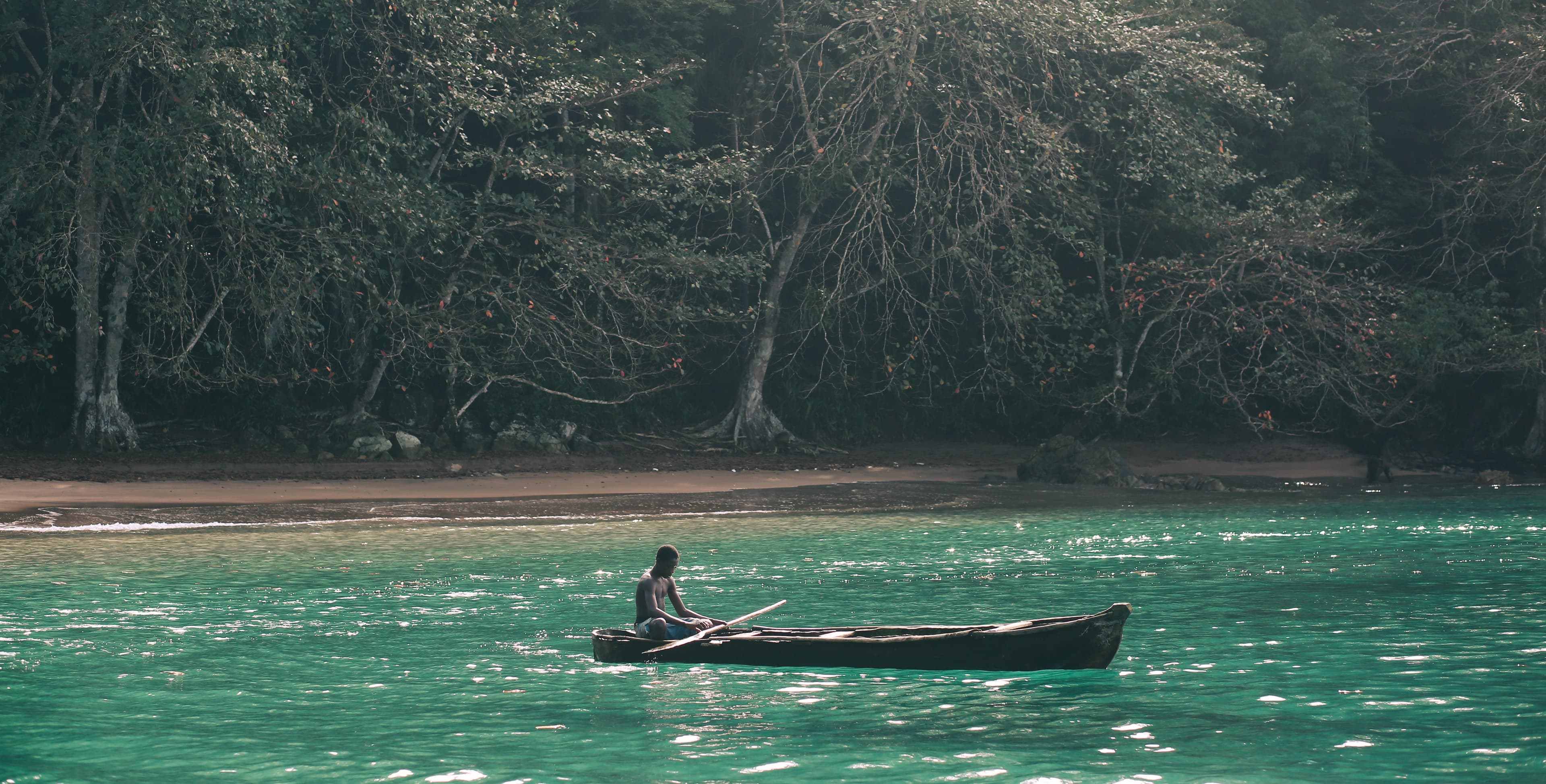 Man sits in a small wooden boat on turquoise water, with a beach and jungle in the background